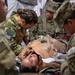 Military medical personnel in camouflage uniforms are attending to a patient lying on a stretcher, performing what appears to be a medical training exercise or field treatment scenario. Multiple soldiers surround the patient, who has a breathing mask/oxygen device on their chest. One soldier's name tape reading 'PUGLIA' is visible on their uniform.