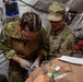 Two female military medical personnel are working together in a field medical tent, performing a medical procedure on a patient or training mannequin. One individual wears Canadian Armed Forces uniform with a Canadian flag patch, while the other wears a US Army uniform with a gold oak leaf insignia (Major rank) and a medical corps patch. The setting appears to be a joint military medical training exercise or field hospital environment.