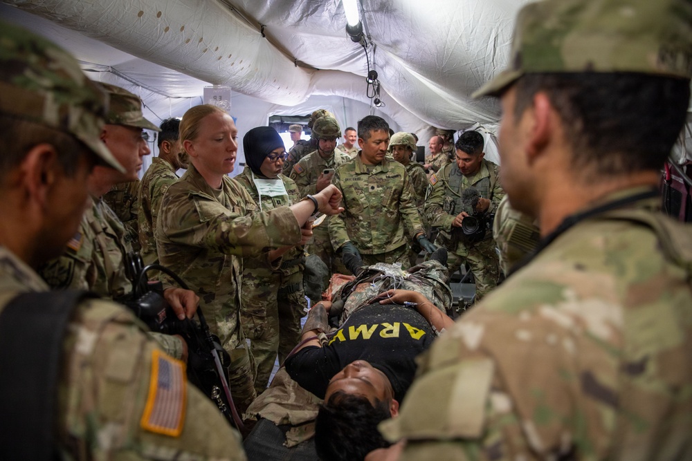 A group of U.S. Army personnel are gathered inside a military field tent conducting what appears to be a medical training exercise or casualty simulation. A person wearing an 'ARMY' shirt is lying on a stretcher or litter while military medical personnel attend to them. Multiple soldiers in OCP (Operational Camouflage Pattern) uniforms surround the patient, with at least one female soldier appearing to lead the demonstration.