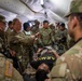 A group of U.S. Army personnel are gathered inside a military field tent conducting what appears to be a medical training exercise or casualty simulation. A person wearing an 'ARMY' shirt is lying on a stretcher or litter while military medical personnel attend to them. Multiple soldiers in OCP (Operational Camouflage Pattern) uniforms surround the patient, with at least one female soldier appearing to lead the demonstration.