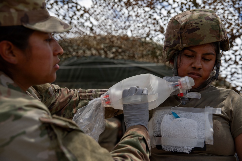 Two military personnel in OCP (Operational Camouflage Pattern) uniforms are engaged in a combat casualty care training exercise. One soldier wearing latex gloves is administering a bag-valve mask (BVM) to another soldier who has simulated chest wound dressings applied. The setting appears to be under a camouflage netting structure, consistent with a field medical training environment.