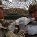Two military personnel in OCP (Operational Camouflage Pattern) uniforms are engaged in a combat casualty care training exercise. One soldier wearing latex gloves is administering a bag-valve mask (BVM) to another soldier who has simulated chest wound dressings applied. The setting appears to be under a camouflage netting structure, consistent with a field medical training environment.