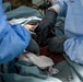 The image depicts a surgical procedure in progress, with multiple medical personnel working on a patient. At least two individuals wearing blue surgical gowns and black nitrile gloves are visible, using surgical instruments including hemostatic clamps/scissors. Surgical drapes, gauze, and visible blood indicate an active operative field.