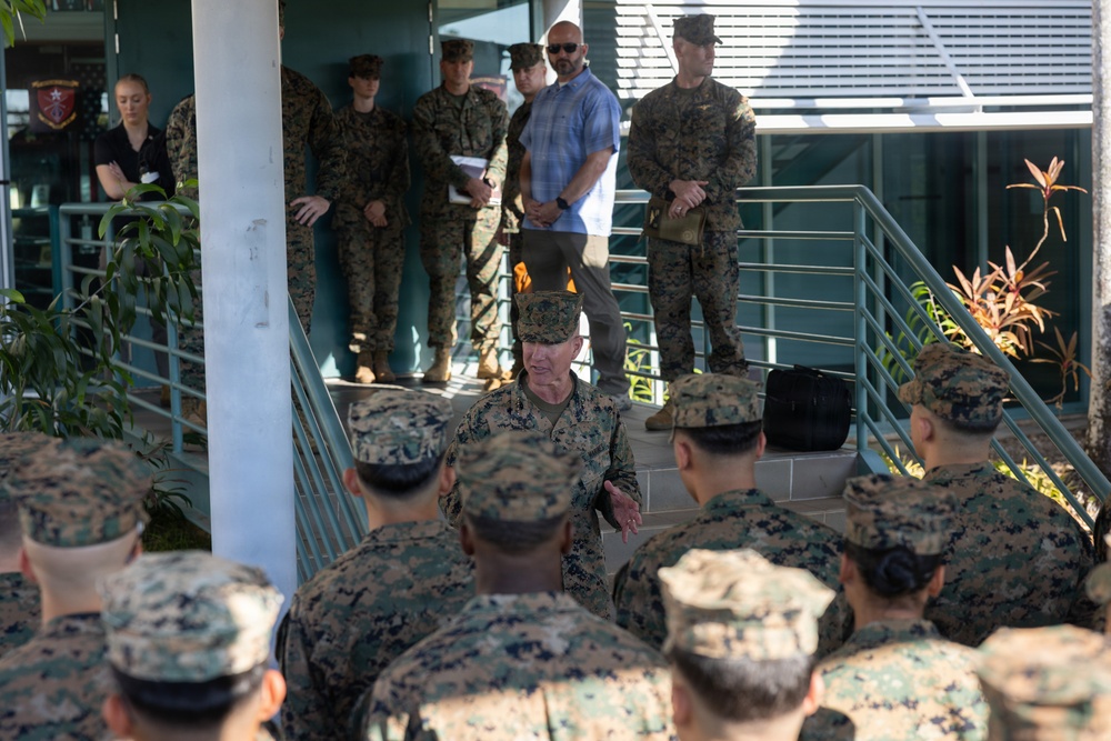 A senior U.S. Marine Corps officer is addressing a group of Marines gathered in front of a military building, likely conducting a briefing or command address. The setting appears to be an outdoor courtyard or entrance area of a military installation, with additional personnel observing from elevated steps in the background. One civilian in a blue shirt is also present among the military personnel.