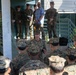 A senior U.S. Marine Corps officer is addressing a group of Marines gathered in front of a military building, likely conducting a briefing or command address. The setting appears to be an outdoor courtyard or entrance area of a military installation, with additional personnel observing from elevated steps in the background. One civilian in a blue shirt is also present among the military personnel.