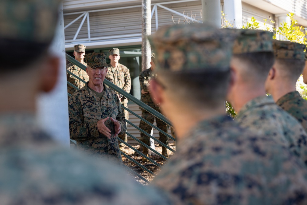 A senior military officer in U.S. Marine Corps digital camouflage (MARPAT) uniform is addressing a group of Marines outdoors near a building with stairs and railings. The central figure appears to be speaking or briefing the assembled personnel, with several Marines listening attentively. The setting appears to be a military installation, likely during daylight hours.