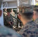 A senior military officer in U.S. Marine Corps digital camouflage (MARPAT) uniform is addressing a group of Marines outdoors near a building with stairs and railings. The central figure appears to be speaking or briefing the assembled personnel, with several Marines listening attentively. The setting appears to be a military installation, likely during daylight hours.