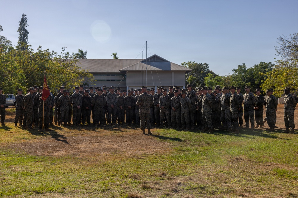 A large formation of military personnel, appearing to be U.S. Marines based on their digital camouflage uniforms (MARPAT), are assembled on a grassy field in front of a low-profile building surrounded by tropical trees. One individual stands facing the group in a position suggesting they are addressing the formation. A red and yellow guidon flag is visible on the left side of the formation.