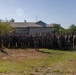 A large formation of military personnel, appearing to be U.S. Marines based on their digital camouflage uniforms (MARPAT), are assembled on a grassy field in front of a low-profile building surrounded by tropical trees. One individual stands facing the group in a position suggesting they are addressing the formation. A red and yellow guidon flag is visible on the left side of the formation.