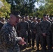 A senior U.S. Marine Corps officer in the foreground is addressing a large formation of Marines in an outdoor setting. The speaker appears to be gesturing with his hands while speaking, suggesting a briefing, command address, or after-action review. The assembled Marines are standing at ease in uniform, attentively facing the speaker.