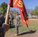A military formation scene showing U.S. Marines in woodland MARPAT camouflage uniforms assembled outdoors. A senior Marine, likely an officer or senior NCO, is addressing the formation with a raised hand gesture. A red guidon flag with yellow markings (partially visible, appearing to show '1/5' or similar unit designation) is held in the foreground.