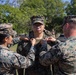Three military personnel in MARPAT (Marine pattern) camouflage uniforms are participating in what appears to be a promotion or award pinning ceremony outdoors. Two service members, one on each side, are simultaneously pinning insignia onto the collar or chest area of the central figure. The ceremony is taking place on a grassy area with trees in the background, with additional personnel visible in the distance.