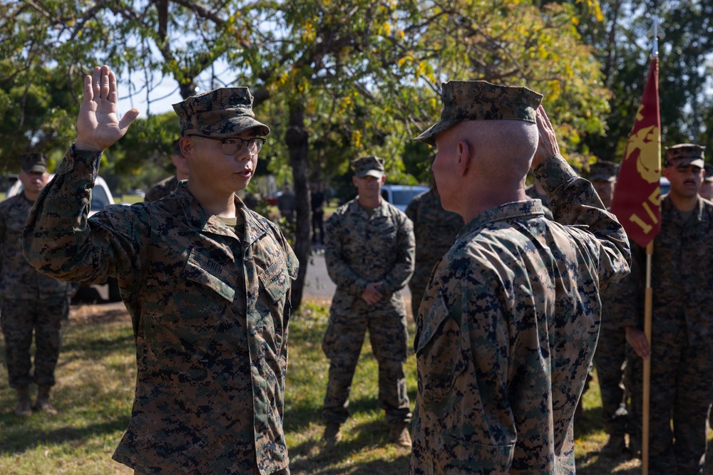 A U.S. Marine Corps ceremony is taking place outdoors, likely a reenlistment or promotion ceremony. A Marine on the left is raising their right hand while a senior Marine facing them renders a salute. Additional Marines in MARPAT digital camouflage uniforms stand in formation in the background, with a Marine Corps guidon visible on the right.