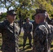 A U.S. Marine Corps ceremony is taking place outdoors, likely a reenlistment or promotion ceremony. A Marine on the left is raising their right hand while a senior Marine facing them renders a salute. Additional Marines in MARPAT digital camouflage uniforms stand in formation in the background, with a Marine Corps guidon visible on the right.