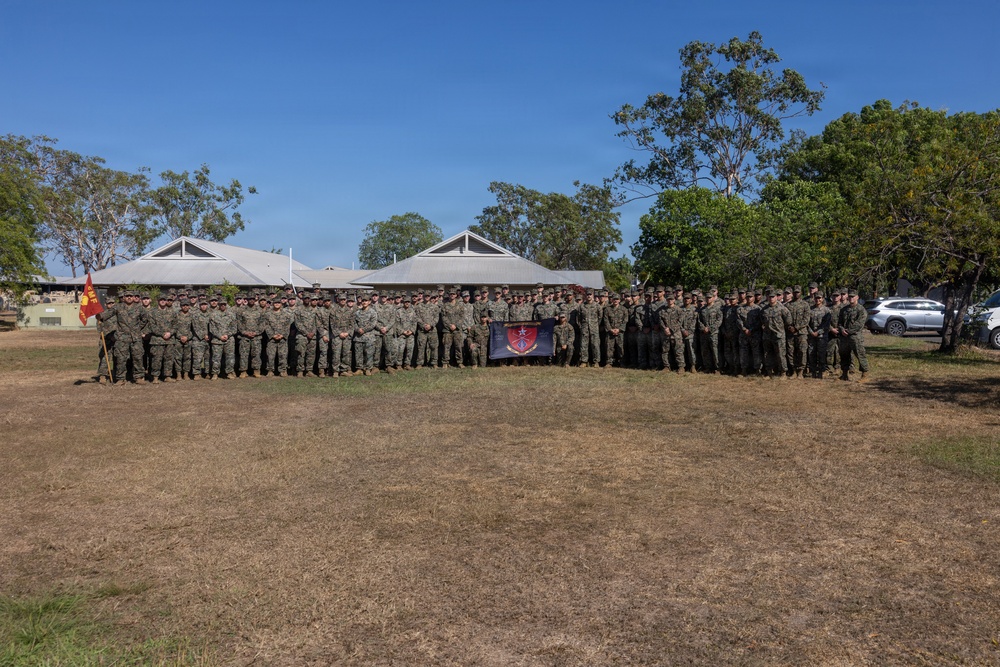 A large group of military personnel, appearing to be U.S. Marines in woodland/digital camouflage uniforms, are posed for a formal group photograph outdoors. They are standing in a single extended row on a dry grass field, with low-profile buildings and eucalyptus trees visible in the background, suggesting a location in Australia. A unit guidon flag and a dark unit banner are visible among the group.