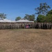 A large group of military personnel, appearing to be U.S. Marines in woodland/digital camouflage uniforms, are posed for a formal group photograph outdoors. They are standing in a single extended row on a dry grass field, with low-profile buildings and eucalyptus trees visible in the background, suggesting a location in Australia. A unit guidon flag and a dark unit banner are visible among the group.