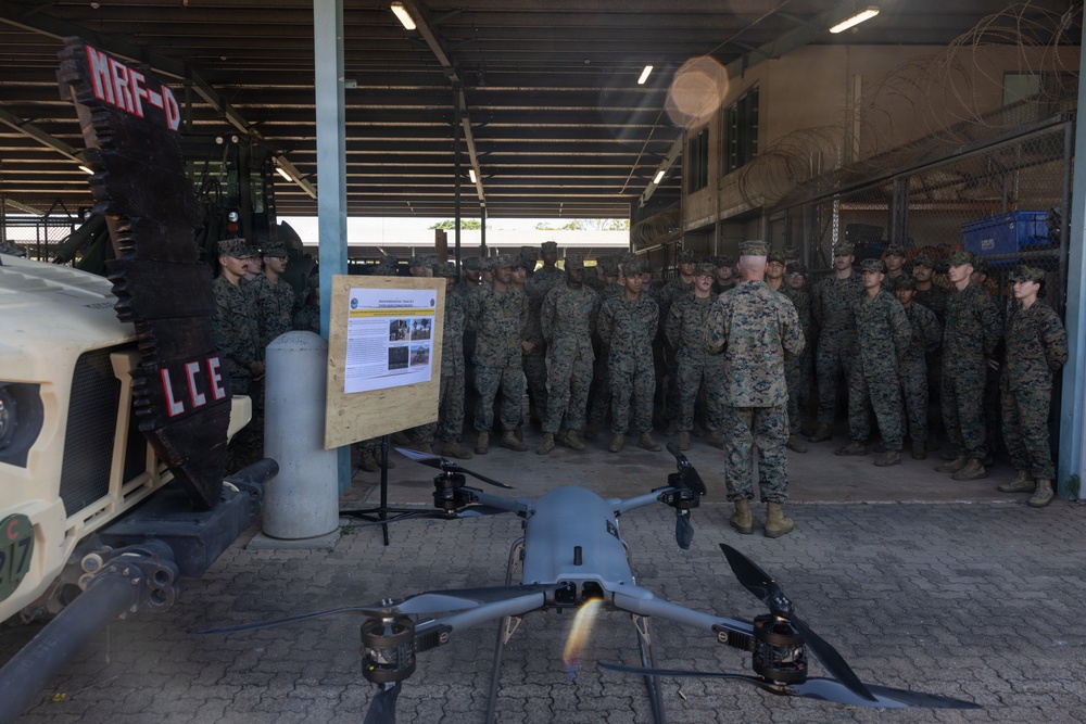 A military briefing is taking place in an outdoor covered facility where a senior military officer addresses a large group of uniformed personnel. A quadcopter drone is displayed in the foreground as part of the briefing, alongside an informational board and a military vehicle. The setting appears to be a forward operating base or military installation, identifiable by the 'MRF-D' (Marine Rotational Force - Darwin) signage.