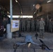 A military briefing is taking place in an outdoor covered facility where a senior military officer addresses a large group of uniformed personnel. A quadcopter drone is displayed in the foreground as part of the briefing, alongside an informational board and a military vehicle. The setting appears to be a forward operating base or military installation, identifiable by the 'MRF-D' (Marine Rotational Force - Darwin) signage.
