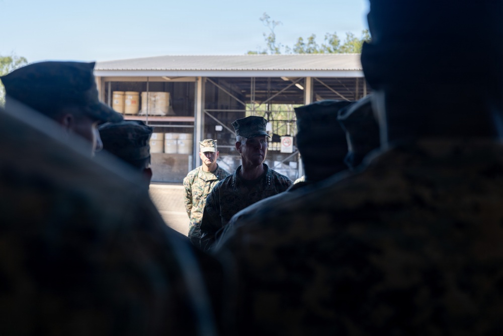 A group of military personnel in camouflage uniforms are gathered in what appears to be a briefing or formation outdoors. Two individuals in the center foreground appear to be addressing or facing the assembled group, with one wearing a distinctive peaked cap. The setting appears to be a military installation or training facility with an open storage structure visible in the background.