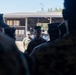 A group of military personnel in camouflage uniforms are gathered in what appears to be a briefing or formation outdoors. Two individuals in the center foreground appear to be addressing or facing the assembled group, with one wearing a distinctive peaked cap. The setting appears to be a military installation or training facility with an open storage structure visible in the background.