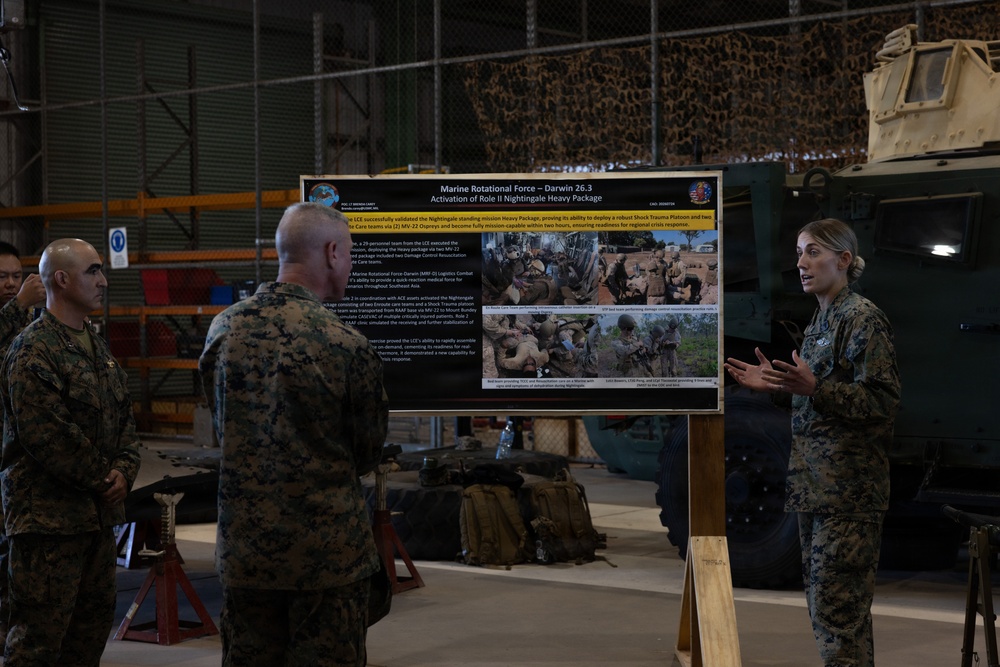 A female Marine in camouflage uniform is briefing a group of fellow Marines in what appears to be a military hangar or maintenance bay. She is presenting in front of a display board titled 'Marine Rotational Force – Darwin 26.3 Activation of Role II Nightingale Heavy Package'. The setting includes military vehicles and equipment visible in the background.