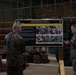 A female Marine in camouflage uniform is briefing a group of fellow Marines in what appears to be a military hangar or maintenance bay. She is presenting in front of a display board titled 'Marine Rotational Force – Darwin 26.3 Activation of Role II Nightingale Heavy Package'. The setting includes military vehicles and equipment visible in the background.