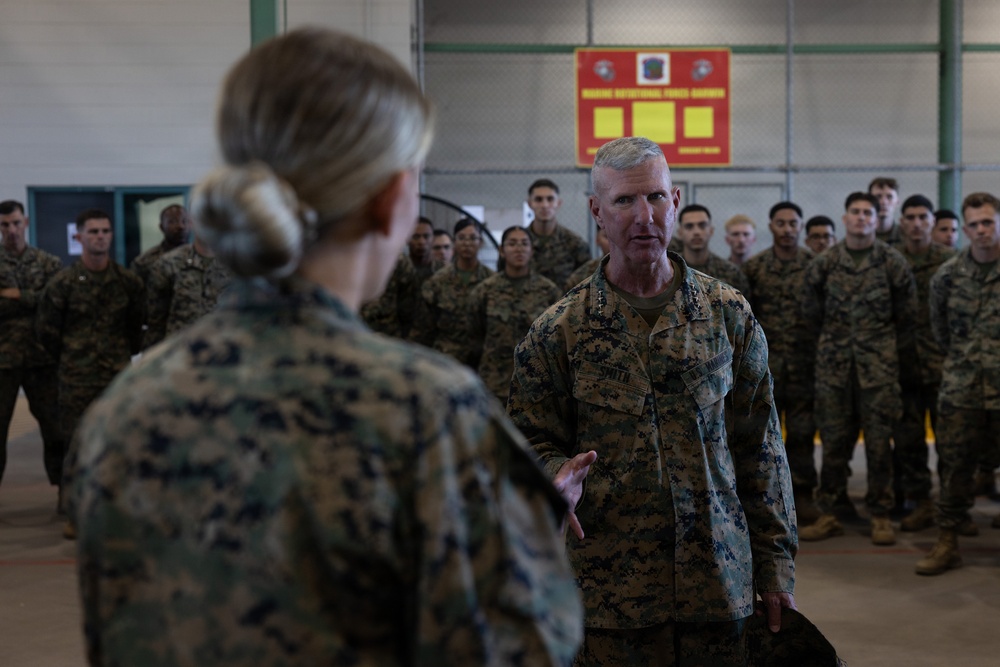 A senior U.S. Marine Corps officer, identifiable by general officer stars on the collar, is addressing a female Marine in the foreground while a large formation of Marines stands at attention in the background. The setting appears to be a military hangar or large indoor facility. A red sign in the background references 'Marine Rotational Force' suggesting this may be a unit visit or inspection.