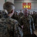 A senior U.S. Marine Corps officer, identifiable by general officer stars on the collar, is addressing a female Marine in the foreground while a large formation of Marines stands at attention in the background. The setting appears to be a military hangar or large indoor facility. A red sign in the background references 'Marine Rotational Force' suggesting this may be a unit visit or inspection.