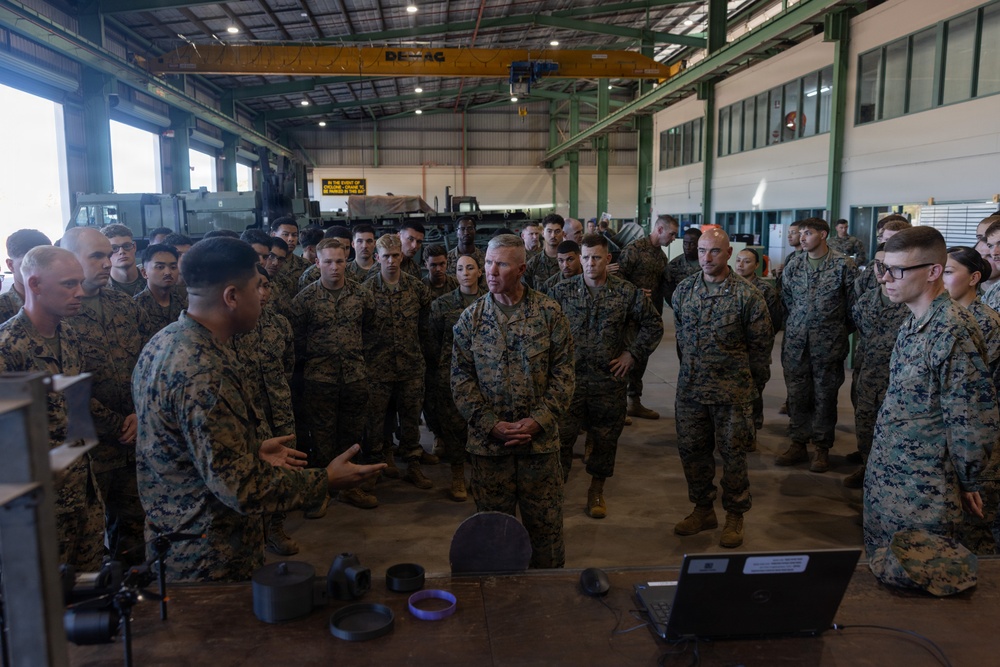 A large group of U.S. Marines in MARPAT camouflage uniforms are gathered inside an industrial hangar or maintenance facility. A Marine appears to be briefing or presenting to the group, including a senior officer, near a table with a laptop and equipment. The setting suggests a unit briefing, demonstration, or inspection event.