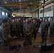 A large group of U.S. Marines in MARPAT camouflage uniforms are gathered inside an industrial hangar or maintenance facility. A Marine appears to be briefing or presenting to the group, including a senior officer, near a table with a laptop and equipment. The setting suggests a unit briefing, demonstration, or inspection event.