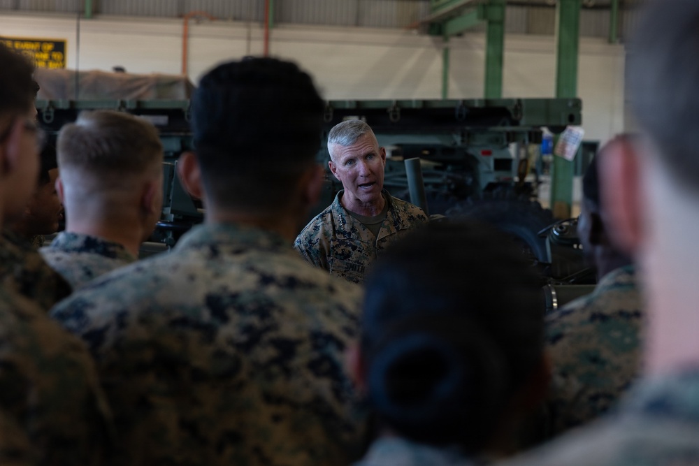 A senior military officer in U.S. Marine Corps MARPAT camouflage uniform is addressing a group of Marines inside what appears to be a military maintenance bay or hangar. The officer, who appears to be of higher rank based on visible collar insignia, is speaking to a gathered group of Marines who have their backs to the camera. Military vehicles or equipment are visible in the background.