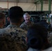 A senior military officer in U.S. Marine Corps MARPAT camouflage uniform is addressing a group of Marines inside what appears to be a military maintenance bay or hangar. The officer, who appears to be of higher rank based on visible collar insignia, is speaking to a gathered group of Marines who have their backs to the camera. Military vehicles or equipment are visible in the background.