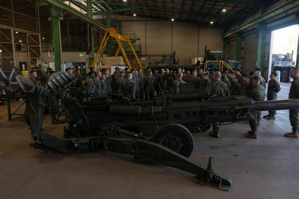 A large group of military personnel in camouflage uniforms are gathered inside an industrial hangar around what appears to be an M777 lightweight howitzer or similar artillery piece. The setting suggests a training or orientation briefing, with an instructor or senior personnel addressing the assembled troops. The facility contains additional military vehicles and equipment visible in the background.