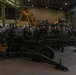 A large group of military personnel in camouflage uniforms are gathered inside an industrial hangar around what appears to be an M777 lightweight howitzer or similar artillery piece. The setting suggests a training or orientation briefing, with an instructor or senior personnel addressing the assembled troops. The facility contains additional military vehicles and equipment visible in the background.