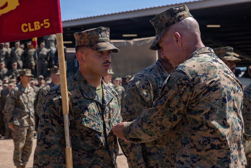 A U.S. Marine Corps ceremony is taking place outdoors, where a senior Marine is pinning a medal or award onto the uniform of a Marine identified by the name tape 'Rodriguez.' A red guidon flag marked 'CLB-5' (Combat Logistics Battalion 5) is visible in the foreground, and a formation of Marines stands in the background.
