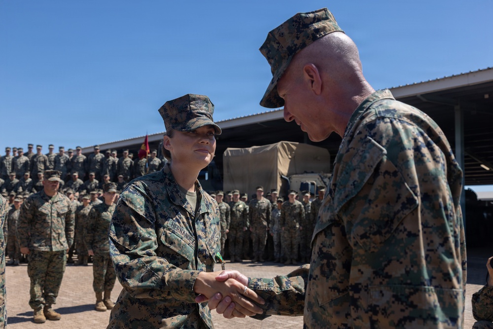 A military award or recognition ceremony is taking place outdoors at what appears to be a U.S. Marine Corps installation. A senior Marine officer is shaking hands with a Marine whose name tape reads 'HERNANDEZ,' likely presenting a commendation or medal. A large formation of Marines in MARPAT camouflage uniforms stands at attention in the background.