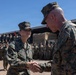 A military award or recognition ceremony is taking place outdoors at what appears to be a U.S. Marine Corps installation. A senior Marine officer is shaking hands with a Marine whose name tape reads 'HERNANDEZ,' likely presenting a commendation or medal. A large formation of Marines in MARPAT camouflage uniforms stands at attention in the background.