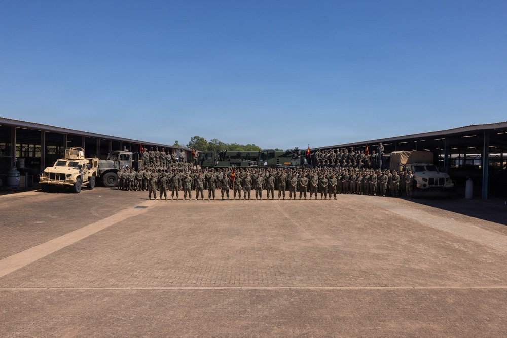 A large formation of military personnel in camouflage uniforms are assembled in a motor pool or vehicle staging area for a group photograph. The formation is arranged in multiple rows across a wide paved courtyard, flanked by covered vehicle bays on both sides. Several military vehicles and unit guidon flags are visible in the background.