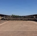 A large formation of military personnel in camouflage uniforms are assembled in a motor pool or vehicle staging area for a group photograph. The formation is arranged in multiple rows across a wide paved courtyard, flanked by covered vehicle bays on both sides. Several military vehicles and unit guidon flags are visible in the background.