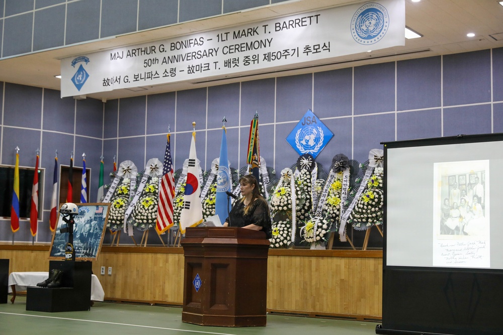 This image shows a 50th Anniversary Ceremony commemorating Major Arthur G. Bonifas and 1st Lieutenant Mark T. Barrett, held at what appears to be a United Nations Command facility in Korea. A woman is speaking at a wooden podium on a stage decorated with numerous floral wreaths and international flags. A projected image of a historical family photograph is visible on a screen to the right.