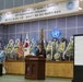 This image shows a 50th Anniversary Ceremony commemorating Major Arthur G. Bonifas and 1st Lieutenant Mark T. Barrett, held at what appears to be a United Nations Command facility in Korea. A woman is speaking at a wooden podium on a stage decorated with numerous floral wreaths and international flags. A projected image of a historical family photograph is visible on a screen to the right.