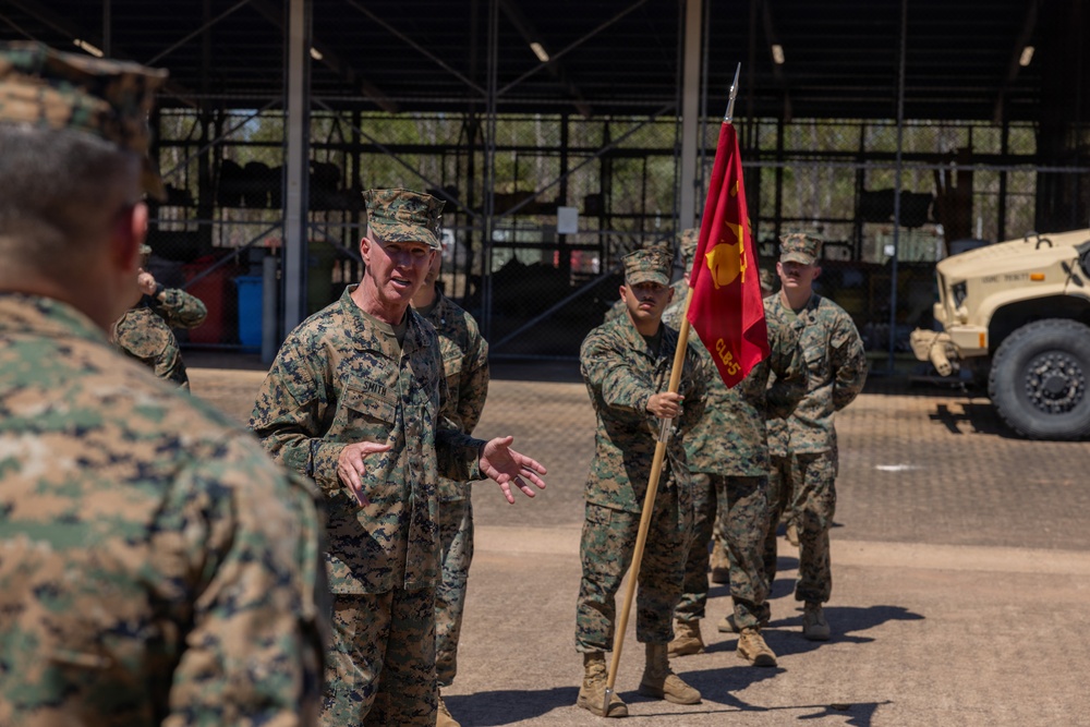 A group of U.S. Marines in MARPAT camouflage uniforms are gathered outdoors at what appears to be a military installation or motor pool area. A senior Marine identified by the name tape 'SMITH' is speaking and gesturing to personnel in front of him, while another Marine holds a red unit guidon flag. The setting includes military vehicles and an open warehouse structure in the background.
