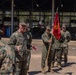 A group of U.S. Marines in MARPAT camouflage uniforms are gathered outdoors at what appears to be a military installation or motor pool area. A senior Marine identified by the name tape 'SMITH' is speaking and gesturing to personnel in front of him, while another Marine holds a red unit guidon flag. The setting includes military vehicles and an open warehouse structure in the background.