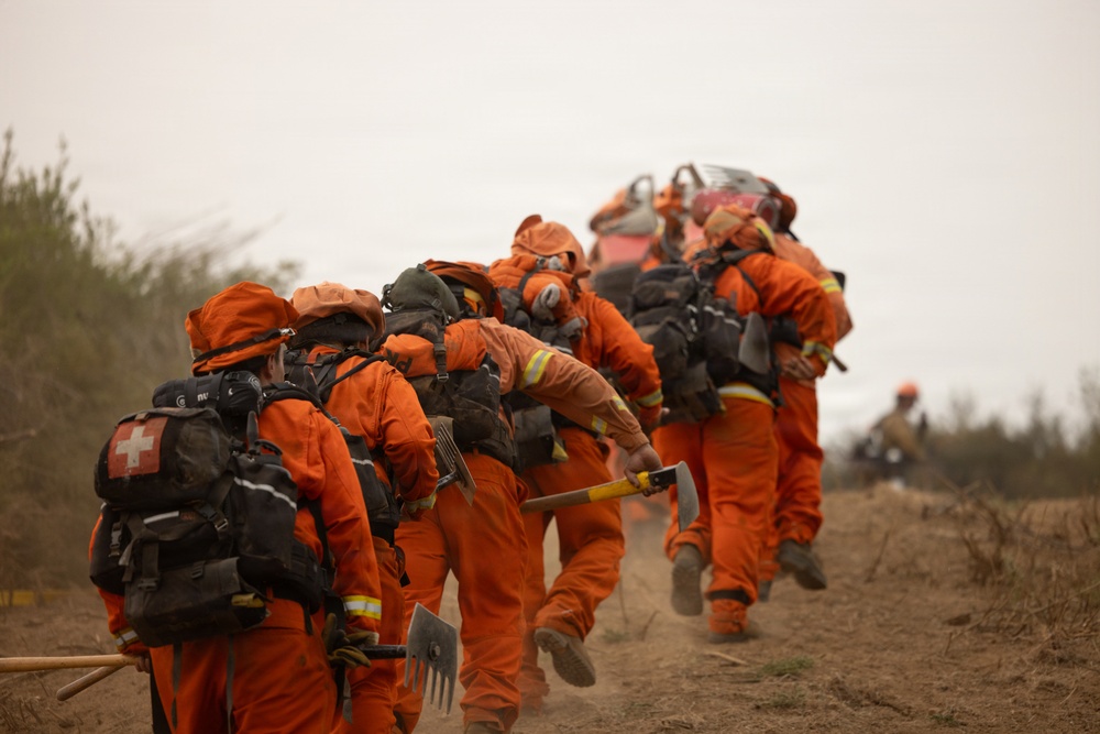 A crew of wildland firefighters dressed in orange Nomex fire-resistant suits are marching in a line along a dirt path, carrying hand tools and equipment. The hazy, smoke-filled sky and dusty terrain suggest they are actively responding to or returning from a wildfire. The group appears to be a hand crew, likely a prison or inmate firefighting crew based on their uniform style.