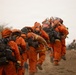 A crew of wildland firefighters dressed in orange Nomex fire-resistant suits are marching in a line along a dirt path, carrying hand tools and equipment. The hazy, smoke-filled sky and dusty terrain suggest they are actively responding to or returning from a wildfire. The group appears to be a hand crew, likely a prison or inmate firefighting crew based on their uniform style.