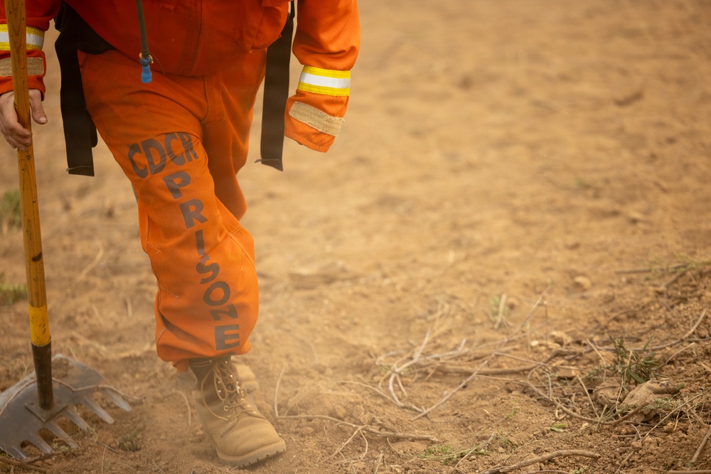 The image shows the lower body of a person wearing an orange jumpsuit marked 'CDCR PRISONER,' indicating they are an incarcerated individual from the California Department of Corrections and Rehabilitation. The person is holding a rake or similar firefighting hand tool and appears to be working on dry, dusty terrain. This is consistent with California's Conservation Camp Program, where incarcerated individuals assist with wildfire suppression and land management.