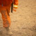 The image shows the lower body of a person wearing an orange jumpsuit marked 'CDCR PRISONER,' indicating they are an incarcerated individual from the California Department of Corrections and Rehabilitation. The person is holding a rake or similar firefighting hand tool and appears to be working on dry, dusty terrain. This is consistent with California's Conservation Camp Program, where incarcerated individuals assist with wildfire suppression and land management.