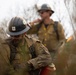 Two U.S. Forest Service wildland firefighters are working in a smoky, vegetation-dense environment. The foreground firefighter wears a dirty hard hat bearing the USFS shield badge and is handling what appears to be a drip torch used for controlled burns. Both personnel are wearing standard yellow Nomex fire-resistant gear that shows significant dirt and soot accumulation.