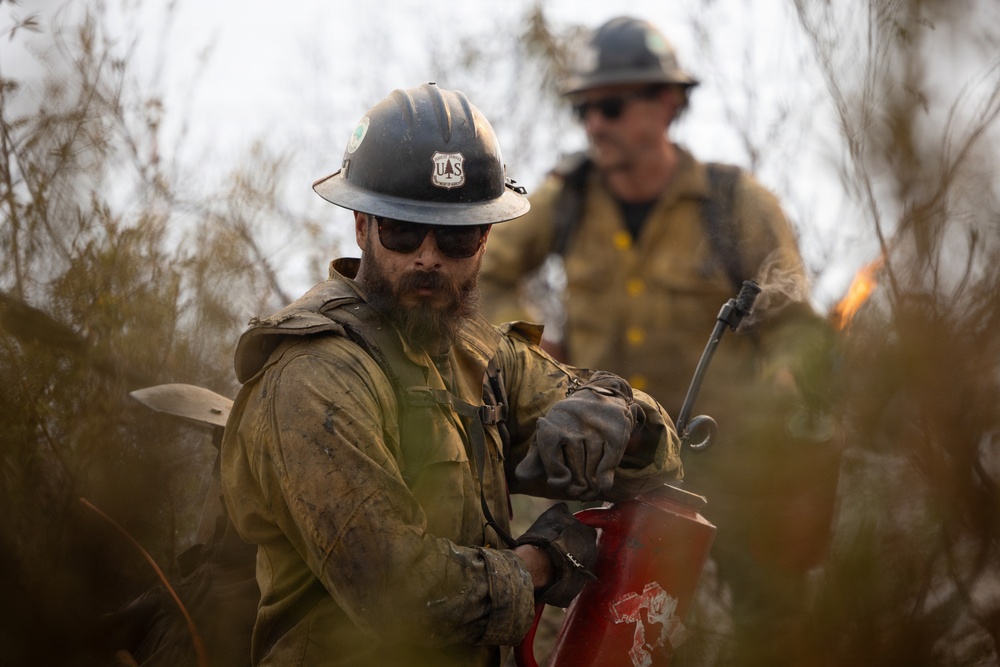 Two U.S. Forest Service wildland firefighters are actively working in a fire environment surrounded by brush and smoke. The foreground firefighter, wearing a USFS hard hat and heavily soiled fire-resistant gear, is operating a red piece of equipment, likely a drip torch or pump. Flames are visible in the background near the second firefighter.