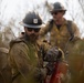 Two U.S. Forest Service wildland firefighters are actively working in a fire environment surrounded by brush and smoke. The foreground firefighter, wearing a USFS hard hat and heavily soiled fire-resistant gear, is operating a red piece of equipment, likely a drip torch or pump. Flames are visible in the background near the second firefighter.